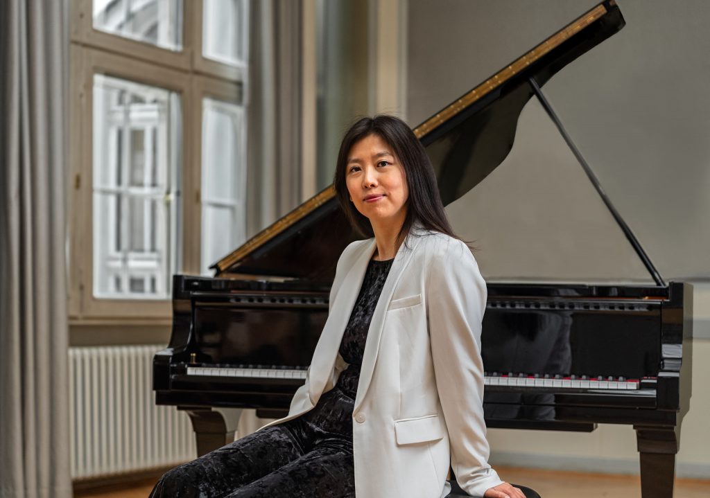 Elegantes Musikerinnenporträt einer asiatischen Pianistin vor einem glänzenden Flügel. Sie trägt einen stilvollen, weißen Blazer und sitzt aufrecht in einem Raum mit einem großen Fenster im Hintergrund. Die weiche Lichtstimmung hebt ihre Gesichtszüge und die Konturen des Instruments harmonisch hervor.
