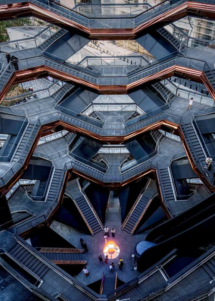 Eine spektakuläre, geometrische Fotografie aus dem Inneren der Skulptur „The Vessel“ in den Hudson Yards, New York. Der Blick ist steil von oben herab direkt in das tief liegende, kreisförmige Zentrum gerichtet. Links und rechts windet sich die komplexe, wabenartige Architektur aus ineinandergreifenden Treppenaufgängen, Plattformen und Schrägen symmetrisch in die Tiefe. Das Zusammenspiel aus dem metallischen Glanz der Oberflächen und den harten, dunklen Schatten in den Zwischenräumen betont die futuristische, fast hypnotische Struktur des Bauwerks.