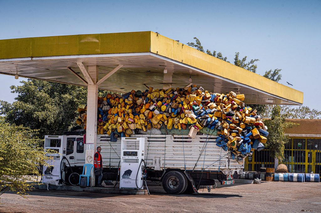 Visuelle Enge an einer Tankstelle: Ein Lkw, beladen mit unzähligen, farbenfrohen Plastikkanistern, passt gerade so unter das Vordach der Station, während die Ladung bedrohlich hoch und breit übersteht.
