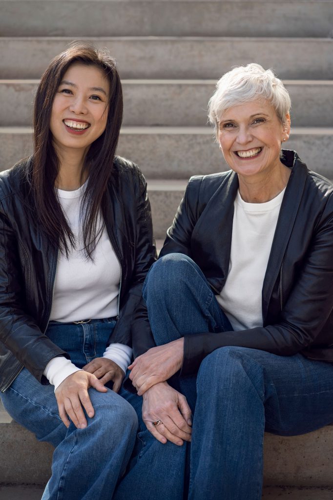 Zwei Frauen – eine junge Asiatin und eine ältere Blondine mit kurzen Haaren – sitzen gut gelaunt auf einer Treppe. Beide sind lässig im Partnerlook mit weißem T-Shirt, Lederjacke und Jeans gekleidet.