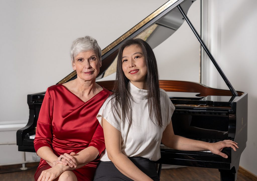 Elegantes Künstlerporträt zweier Künstlerinnen, die nebeneinander vor einem Steinway-Flügel sitzen. Rechts sitzt eine junge asiatische Frau in einer weißen Seidenbluse. Neben ihr eine ältere, blonde Frau in einem festlichen, roten Abendkleid.