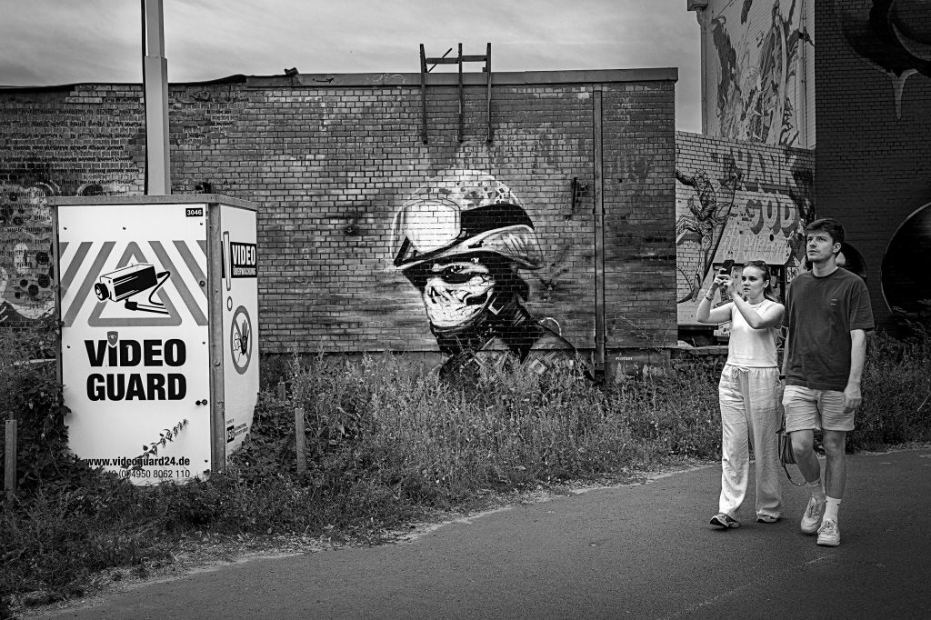 Eine kontrastreiche, monochrome Streetphotography-Aufnahme aus dem Zentrum Berlins. Der Blick fällt auf eine raue Betonmauer, die von einem großflächigen Graffiti dominiert wird. Es zeigt den Kopf eines Mannes, der einen Helm trägt und dessen Mundpartie maskiert ist. Am rechten Bildrand steht ein Passanten-Pärchen, beide halten Kameras hoch und fotografieren die Szene. Auf der linken Seite, leicht im Vordergrund, ist ein technischer Kasten an der Wand befestigt. Auf ihm sind ein grafisches Symbol für eine Videokamera und der deutlich lesbare Schriftzug „Video Guard“ zu sehen.
