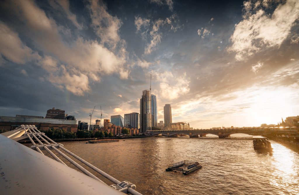 Farbfotografie bei dramatischem Abendhimmel in London: Blick auf die Millennium Bridge über der Themse mit Booten, flankiert von der beleuchteten Tate Modern am linken Ufer.