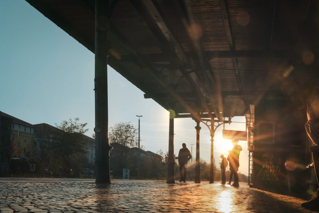 Atmosphärisches Streetphotography-Foto auf dem Bahnsteig der Berliner S-Bahn-Station Lichterfelde West im warmen Herbstlicht. Auf den Gleisen stehen wartende Fahrgäste Die tiefstehende Herbstsonne bricht sich perfekt an der Dachkonstruktion des Bahnsteigs und erzeugt einen leuchtenden, goldenen Sonnenstern.