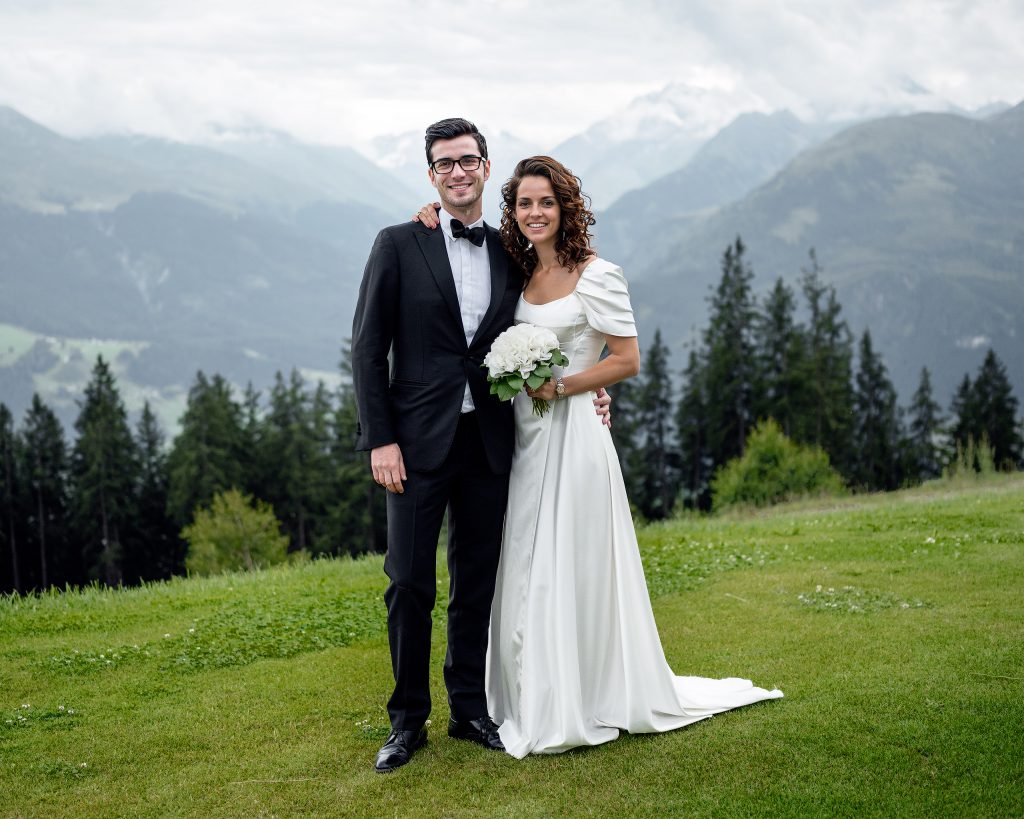 Hochzeitsfotografie eines dunkelhaarigen Brautpaares vor einer beeindruckenden alpinen Bergkulisse: Die Braut im eleganten langen weißen Hochzeitskleid und der Bräutigam mit Brille im klassischen Smoking posieren vor dem Panorama der Alpen.