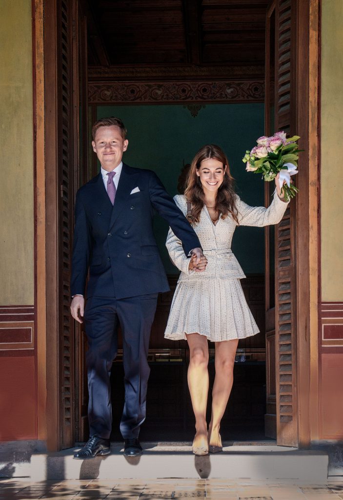 Emotionaler Hochzeitsmoment: Das Brautpaar verlässt Hand in Hand das Standesamt, während die Braut glücklich den Blumenstrauß in die Luft reckt; der dunkle Hintergrund im Türrahmen lässt das Paar strahlen.