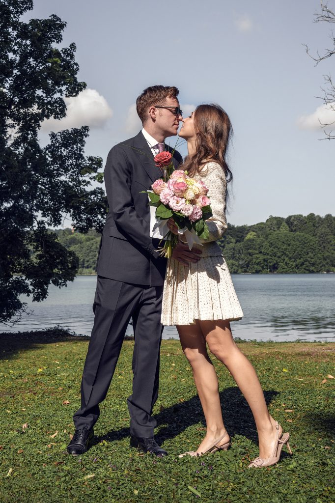 Hochzeitsfotografie am Seeufer: Ein frisch vermähltes Hochzeitspaar im modernen Standesamt-Outfit küsst sich innig vor der malerischen Kulisse des ruhigen Wassers.