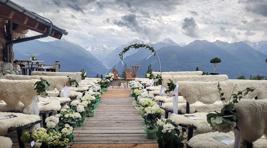 Eventfotografie einer alpinen Outdoor-Hochzeitslocation vor majestätischer Bergkulisse: Eine Allee aus hölzernen Bänken, dekoriert mit weißen Hortensien und gemütlichen Schaffellen, führt perspektivisch auf einen prachtvollen Hochzeitsbogen zu.