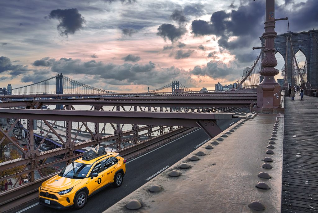 Stimmungsvolle Weitwinkelaufnahme aus einer erhöhten Perspektive von der Brooklyn Bridge hinab auf die darunterliegende Straßenebene. Auf der linken Seite zieht das charakteristische, leuchtend gelbe New Yorker Taxi als farbiger Blickfang seine Spur über den Asphalt. Zur rechten Seite hin ragt die monumentale, hölzerne und stählerne Konstruktion der Brooklyn Bridge mit ihren dicken Tragkabeln und dem Fußgängerweg elegant ins Bild. Über der gesamten Szenerie wölbt sich ein dramatischer Abendhimmel mit tiefen, kontrastreichen Wolkenformationen, die das warme Licht der untergehenden Sonne reflektieren.