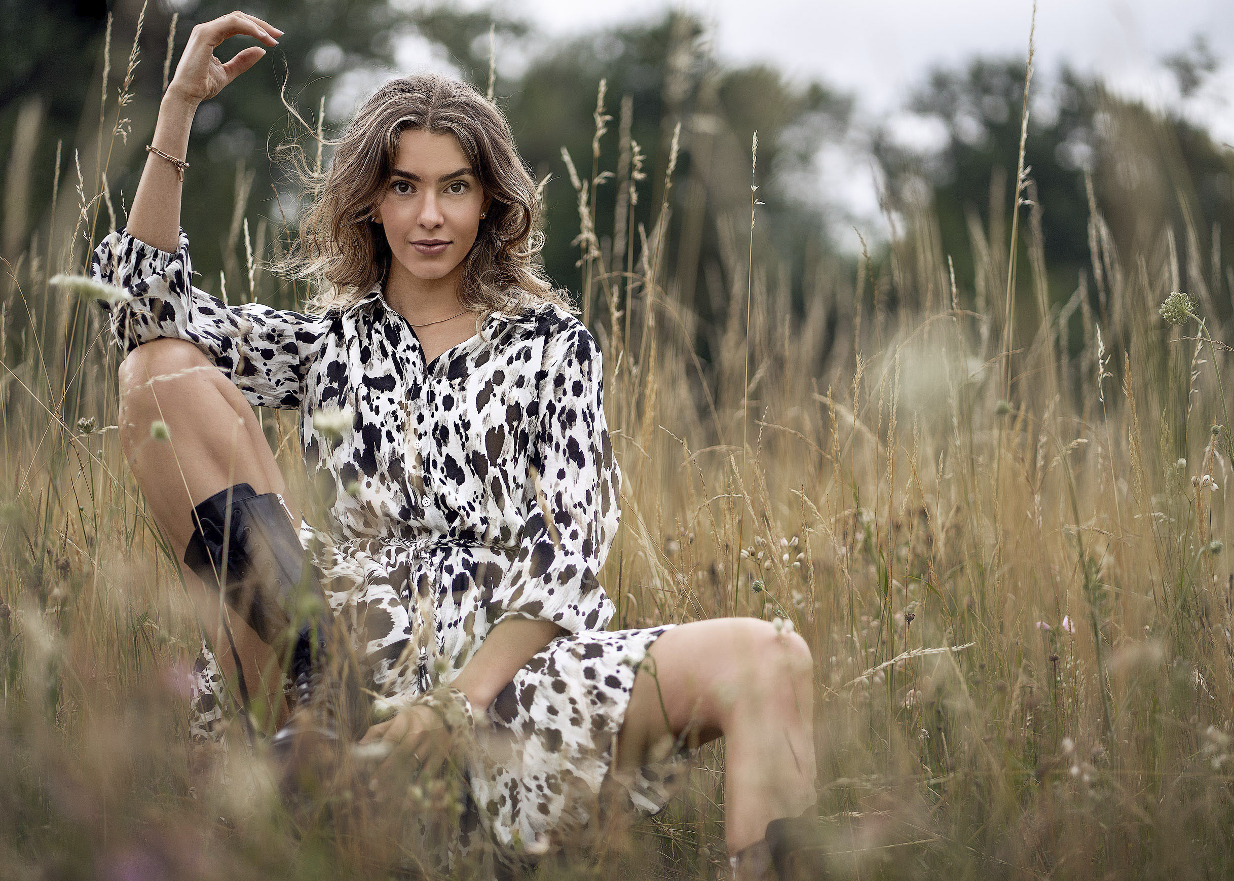 Verträumtes Outdoor-Modefoto, aufgenommen durch unscharfe, hohe Halme einer spätsommerlichen, gelblichen Wiese. Im Fokus steht ein Modell in einem Sommerkleid mit markantem, schwarzem Fleckendruck. Dazu trägt sie als derben Stilbruch dunkle, klobige Chunky Boots. Die verschwommene Perspektive im Vordergrund verleiht dem Bild Tiefe und eine geheimnisvolle Stimmung.