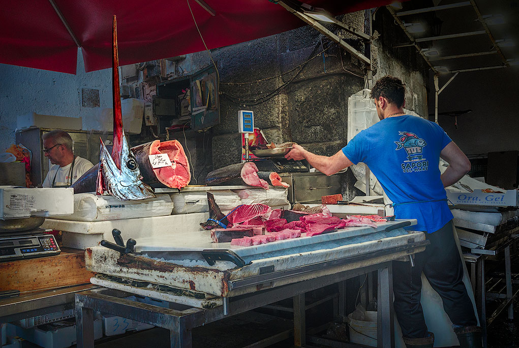Farbfoto von einem Fischstand in der Markthalle von Catania. Vorwiegend in Blaul/Rot Tönen gehalten. Im Vordergrund vor einem Verkaufsstand ein Verkäufe und eine uslage mit Fisch. Besonders hervortretend ist ein großer Schwertfischkopf