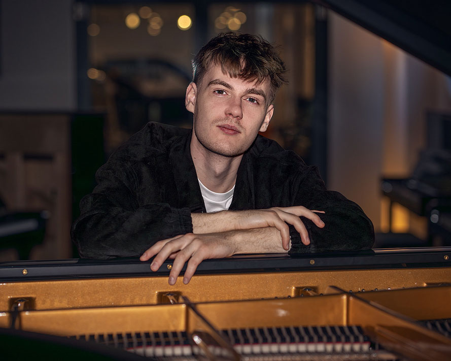 Der Pianist Janik Chêch aufgenommen vor einem Bechstein Flügel, er schaut durch die offene Flügeldecke vor dunklem Hintergrund.