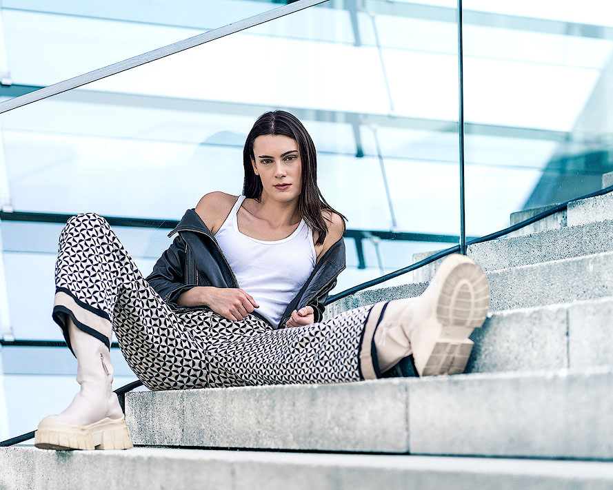 Mode-Shooting vor moderner Glasfassade im kühlen Farbton. Dunkelhaariges Model posiert provokant auf einer Treppe in einer Kombination aus schwarzer Lederjacke, Hose mit grafischem Muster, weißem Top und derben Boots.