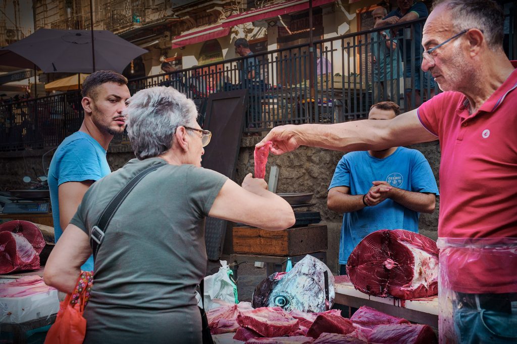 Farbfoto von einem Fischstand auf dem Fischmarkt von Catania. Vorwiegend in Blau/Rot Tönen gehalten. Im Vordergrund sieht man einen Verkäufer an einem Thunfischstand der einer Verkäuferin dein Stück Thunfisch zur Begutachtung hinhält.