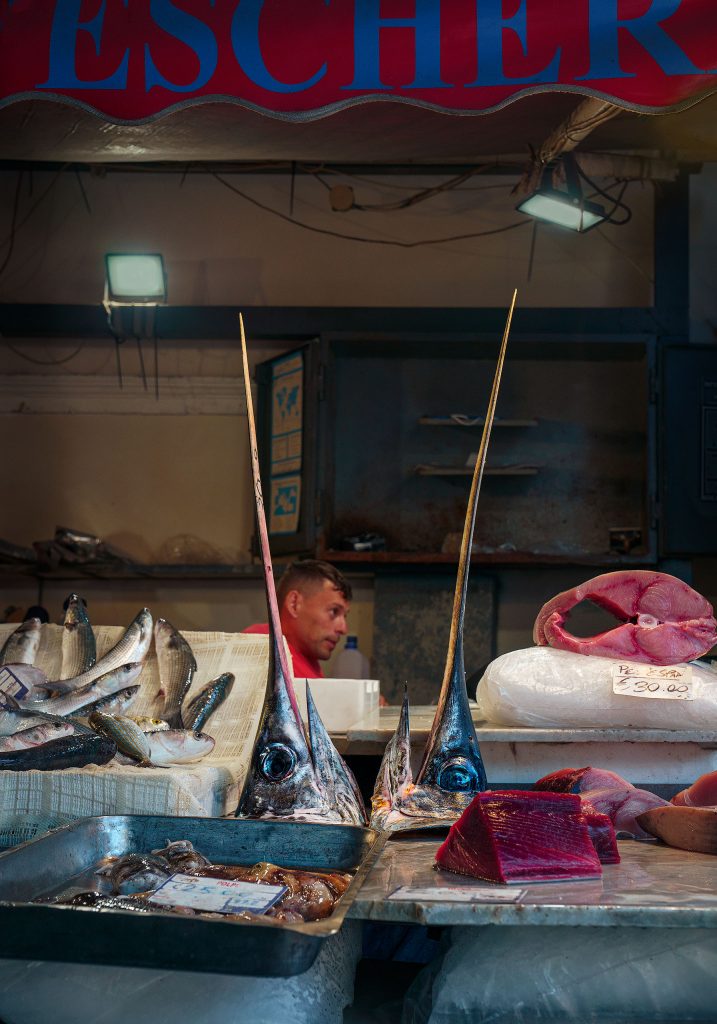 Farbfoto von einem Fischstand in der Markthalle von Catania. Vorwiegend in Petrol/Rot Tönen gehalten. Im Vordergrund sind 2 große Schwertfischköpfe auf einem Verkaufsstand aufgebahrt. Im Hintergrund der Kopf einses Verkäufers.