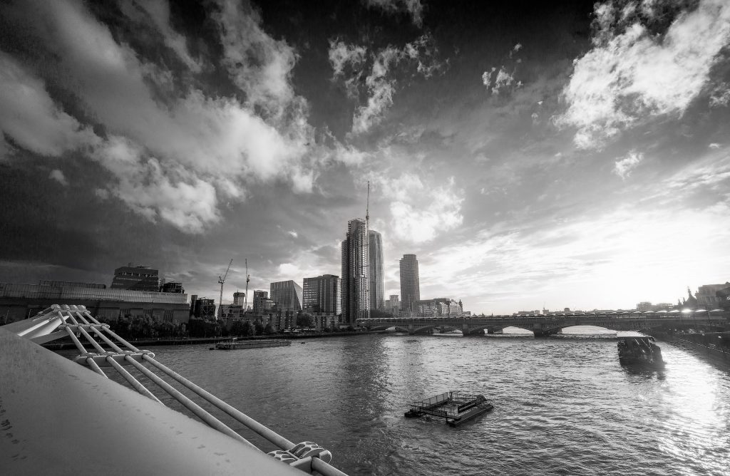 Schwarz/ Weißfotografie bei dramatischem Abendhimmel in London: Blick auf die Millennium Bridge über der Themse mit Booten, flankiert von der beleuchteten Tate Modern am linken Ufer.