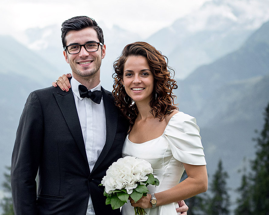 Junges Hochzeitspaar vor Bergkulisse. Sie trägt ein weißes langes Hochzeitkleid, er einen Smoking.
