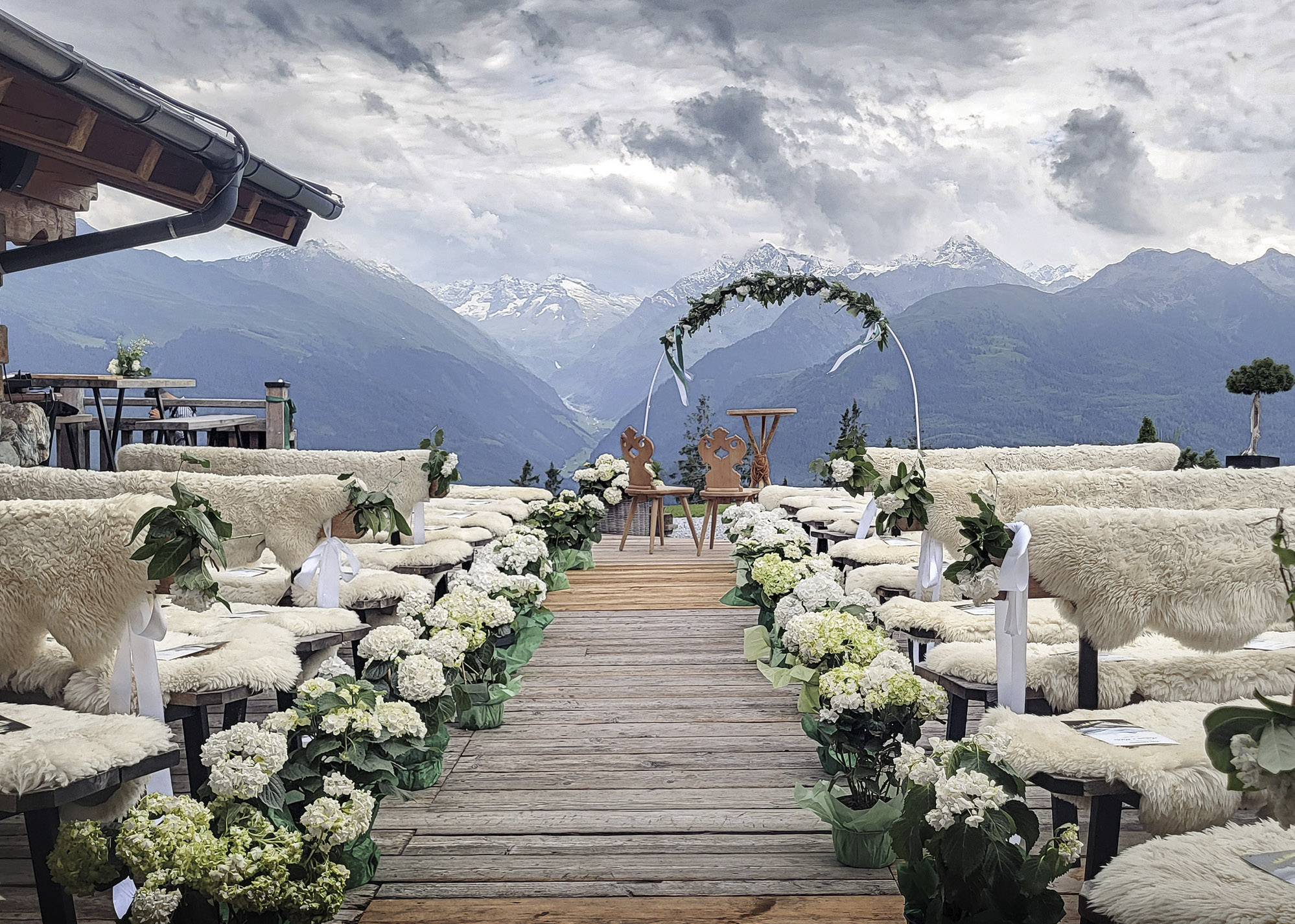 Eventfotografie einer alpinen Outdoor-Hochzeitslocation vor majestätischer Bergkulisse: Eine Allee aus Bänken, dekoriert mit weißen Hortensien und gemütlichen Schaffellen, führt perspektivisch auf einen prachtvollen Hochzeitsbogen zu.