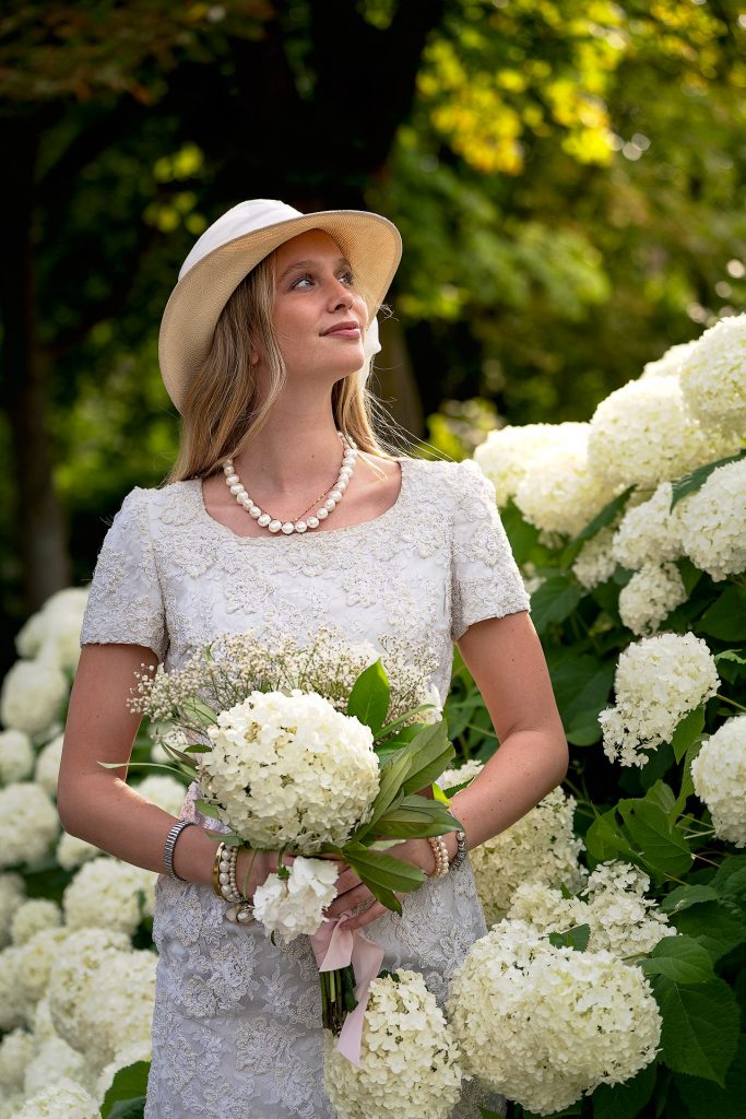 Das Foto einer jungen, blonden Frau in einem weißen Spitzenkleid, einer Braut. Sie trägt einen Hut und hat einen Blumenstrauss in der Hand. Sie steht vor einer Blumenhecke.
