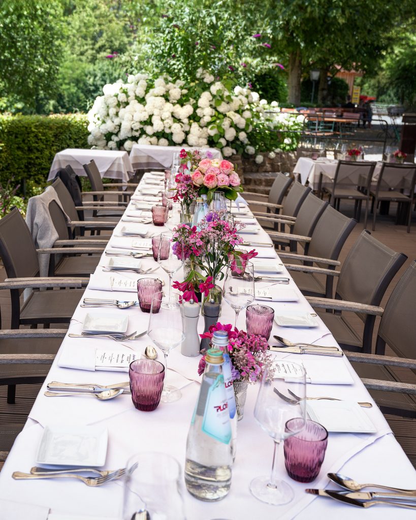 Foto einer festlich gedeckten Tafel, die perspektivisch auf den Fluchtpunkt in der Mitte zuläuft. Rosa Blüten und Christall Gläser geben einen Farbakzent. Im Hintergrunde eine Hecke aus weißen Hortensien.