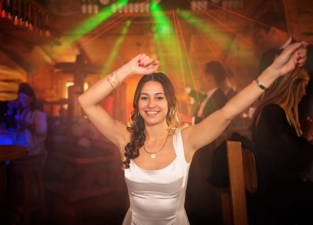 Foto einer jungen, brünetten Frau mit langen locken. Sie lacht in die Kamera, hat die Arme weit ausgestreckt und trägt eine weißes, abendliches Kleid. Im Hintergrund eien Partyszene.