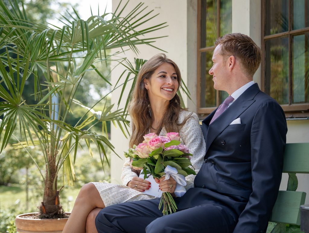 Ein junges Hochzeitspaar auf einer Bank. Sie strahlt ihn verliebt and er schaut ihr in die Augen. In den Händen hält sie einen Rosenstrauss. Im Hinetergrund eine Palme.