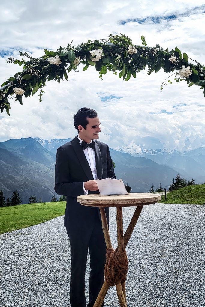 Ein junger Mann im Smoking vor einer beeindruckenden Bergkulisse. Er steht an einem Stehpult und hält ein Schriftstück, für seine Hochzeitsrede in der Hand. Über ihm eine Blumengirlande mit weißen Blüten.