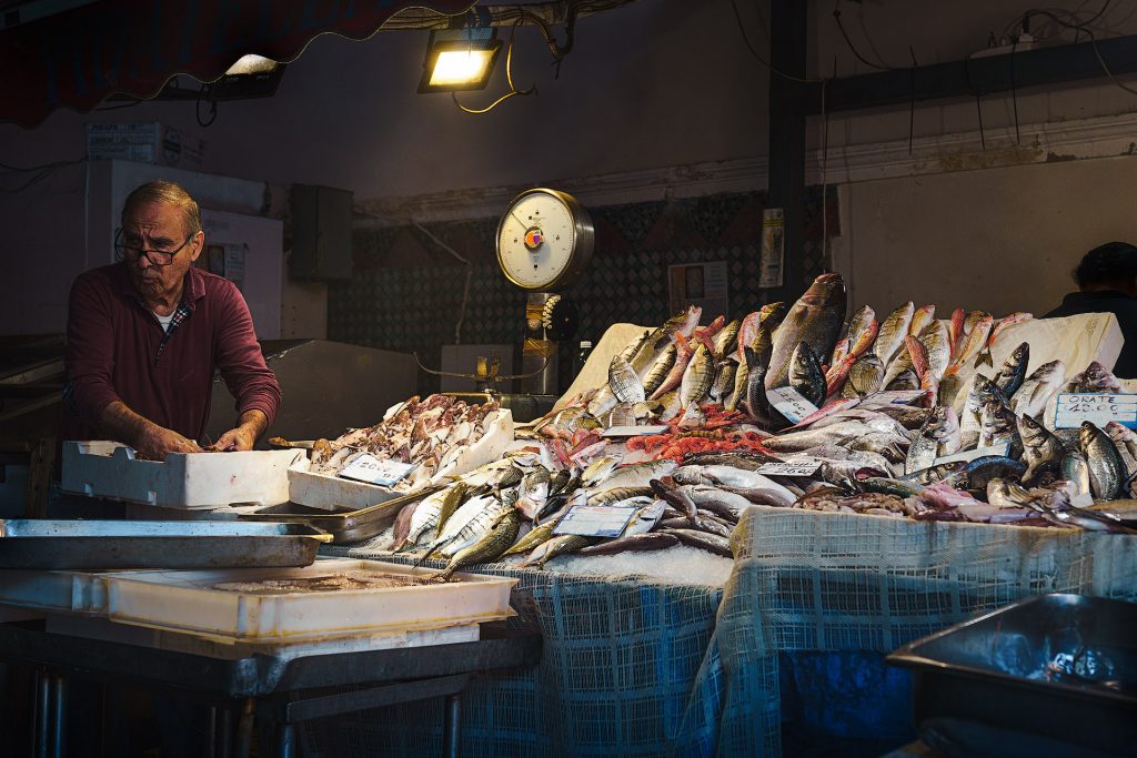 Auf der linken Seite des Bildes steht ein älterer Fischverkäufer mit Brille. Seine Hände stecken in einer Styroporbox auf einem Marktstand, der überreich mit seiner Ware, frischem Fisch, gefüllt ist. Die Szene spielt in einem dunklen Innenraum, wobei der Marktstand hell beleuchtet ist und den Fokus auf den Verkäufer und den Fisch legt.