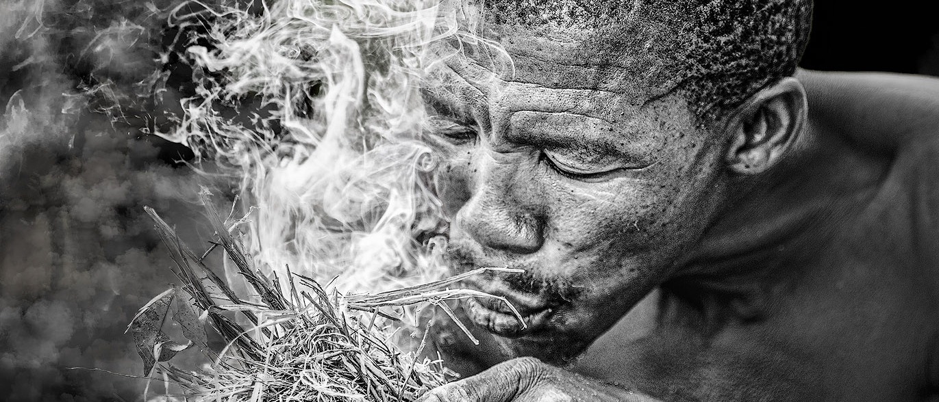 Fine-Art-Fotografie in Schwarz-Weiß, auf dem Foto ist der Kopf eines Mannes zu sehen, der ein brennendes Stroh Büschel in der Hand hält und dieses Feuer durch Blasen verstärkt.