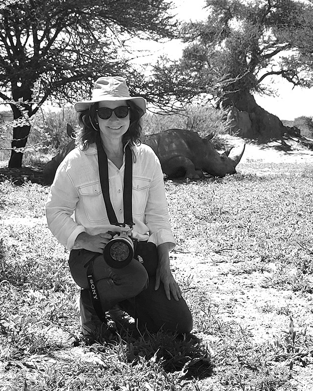 Die Fotografin Barbara Westphalen hockt im Vordergrund mit einer Kamera in der Hand auf einem kargen Untergrund, der mit struppigem Gras bewachsen ist. Direkt hinter ihr liegt ein schlafendes Rhinozeros. In der Umgebung sind einige dürre Akazien zu sehen.
