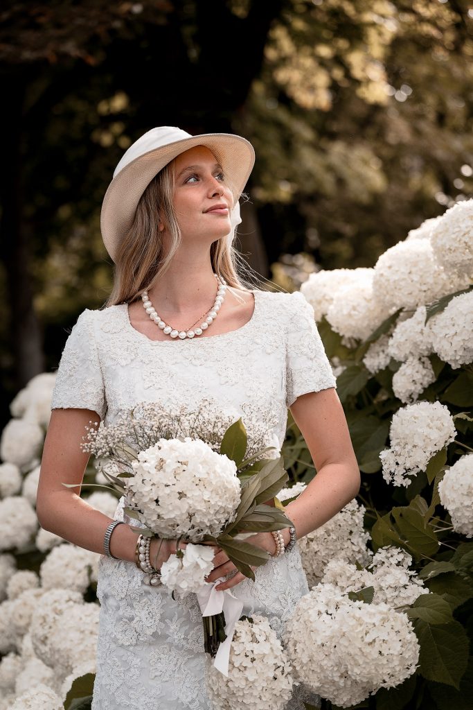 Das Foto einer jungen, blonden Frau in einem weißen Spitzenkleid, einer Braut. Sie trägt einen Hut und hat einen Blumenstrauß in der Hand. Sie steht vor einer weißen Hortensienhecke.