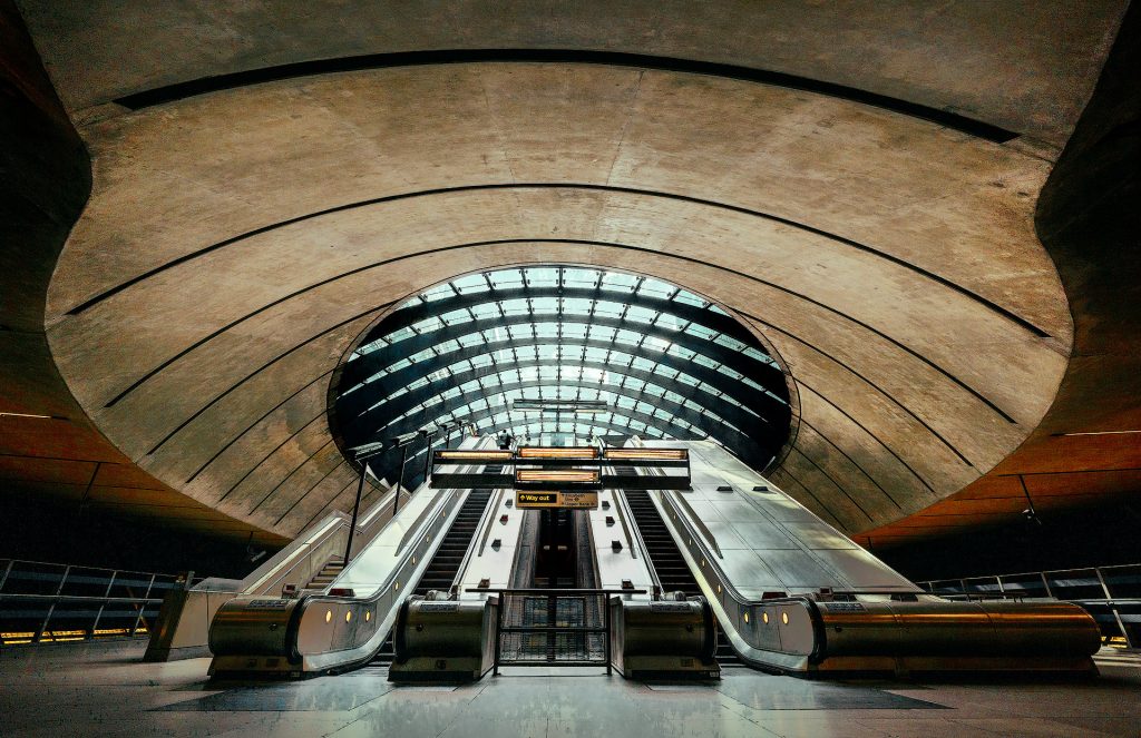 Weitwinkelaufnahme aus dem Inneren der U-Bahn-Station Canary Wharf in London. Die beeindruckende moderne Architektur wird durch ein hohes, rundes Glasdach dominiert, das helles Tageslicht in den Raum lässt. Breite Rolltreppen führen aus der Tiefe nach oben in Richtung des Lichts. Am oberen Ende einer Rolltreppe ist eine einzelne Person klein zu erkennen, was die monumentale Weite der architektonischen Konstruktion unterstreicht.