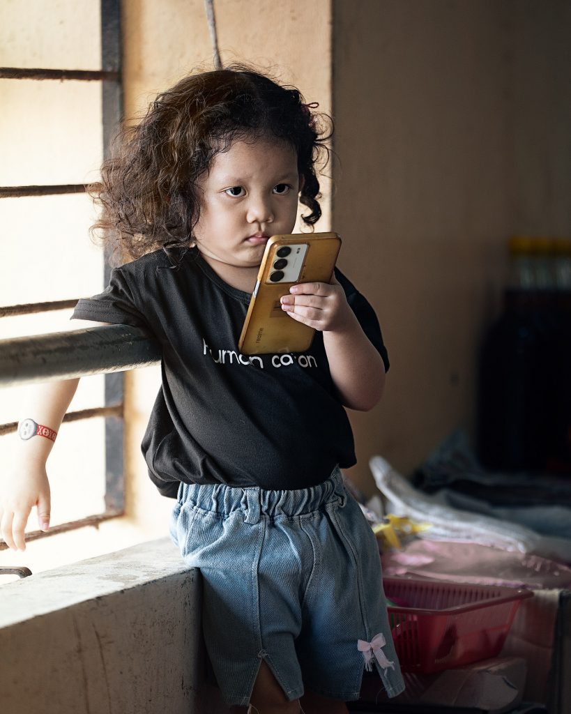 Halbseiten-Porträt eines kleinen asiatischen Mädchens mit strubbeligen Locken und einem schmollenden Gesicht. Sie trägt ein schwarzes T-Shirt und Jeansbermudas, hat markante Knopfaugen und ist bis zum Oberschenkel zu sehen. Sie steht vor einem Fenster, während von links das Licht durch ein Fenster mit Gitterstäben einfällt.