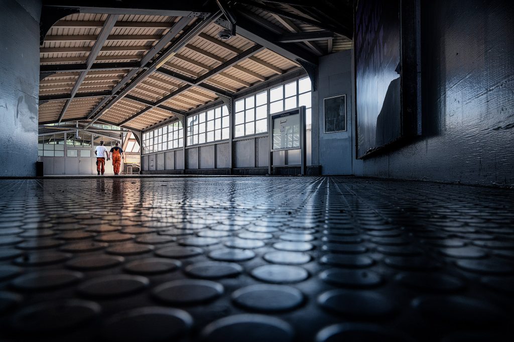 Innenaufnahme der Eingangshalle des S-Bahnhofs Westkreuz aus niedriger Perspektive, die den architektonischen Raum betont. Der größte Teil des Bildes wird vom grauen Noppenboden eingenommen. Die Halle ist von einer Dachkonstruktion aus Metall überdacht. Rechts erstreckt sich eine Fensterfront entlang des Raumes. Am Ende der Halle gehen zwei Männer in orangefarbener Arbeitskleidung.