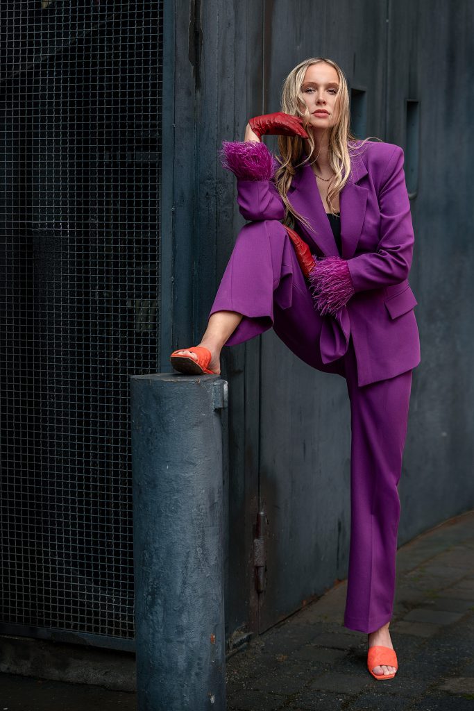 Ein hellblondes, langhaariges Model posiert in einem industriellen Umfeld vor einem dunklen Lastenaufzug, links ist ein Gitter zu sehen. Sie steht provokativ mit einem orangefarbenen Pantoletten-beschuhten Fuß auf einer halbhohen Metallsäule. Sie trägt einen auffälligen, leuchtend lilafarbenen Hosenanzug mit Federbesatz an den Ärmelmanschetten und dazu kontrastierende rote Handschuhe. Der Hintergrund ist dunkel, und kühl gehalten, was den extravaganten Look hervorhebt.