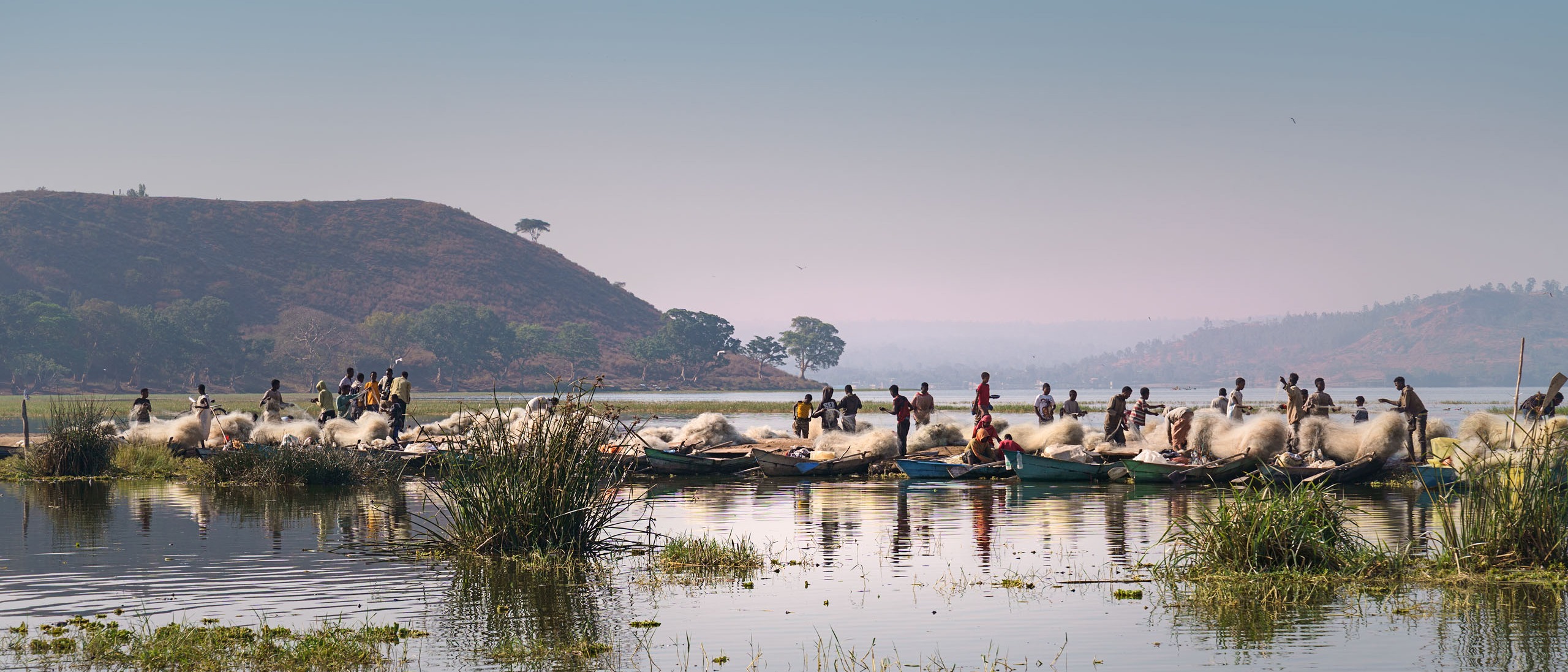 Auf dem Awassa-See sind im Hintergrund mehrere Boote und große Mengen von Fischernetzen zu sehen, die im Morgenlicht glänzen. Im Vordergrund erstreckt sich die Wasserfläche des Sees, dahinter zeichnen sich im Dunst Berge ab.