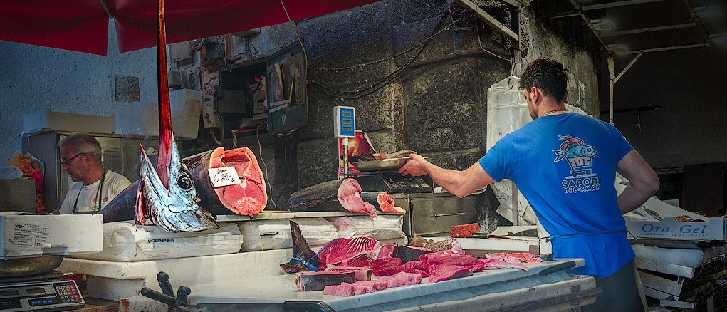 Farbfoto von einem Fischstand in der Markthalle von Catania. Vorwiegend in Blau-l/Rottönen gehalten. Im Vordergrund vor einem Verkaufsstand ein Verkäufer und eine Auslage mit Fisch. Besonders hervortretend ist ein großer Schwertfischkopf.