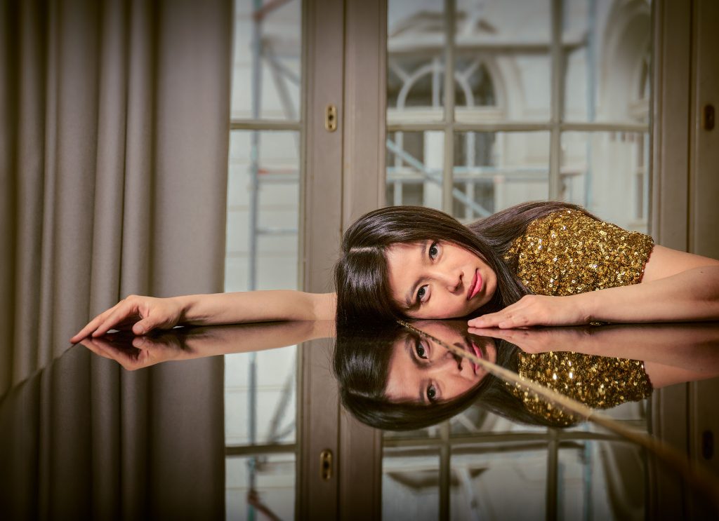 Porträt der Pianistin Huijing Han: Sie liegt mit dem Oberkörper seitlich ausgestreckt auf ihrem rechten Arm, der auf einem Flügel ruht. Sie trägt eine goldene Paillettenjacke. Der Flügel spiegelt sie sowie ein Fenster im Hintergrund wider.