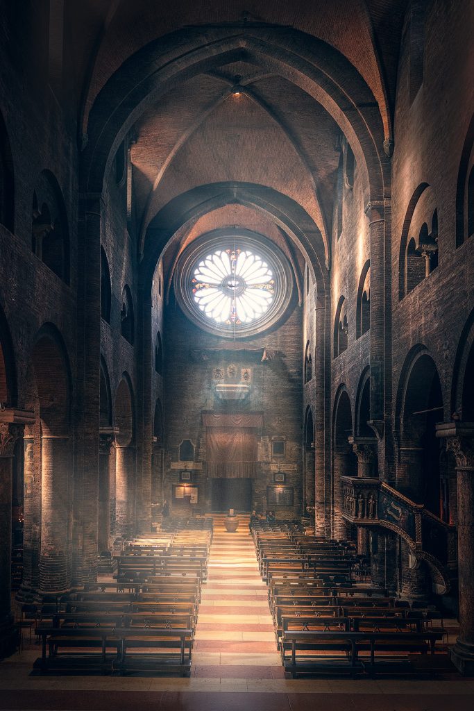 Dunkle und geheimnisvolle Fine-Art-Aufnahme des Inneren einer der Kathedrale in Modenea, mit einem zentralen Blick auf die Rosette im Hauptschiff. Von links und von der Rosette fallen stimmungsvolle Lichtstrahlen in den Innenraum, der zu beiden Seiten von Kirchenbänken gesäumt wird.
