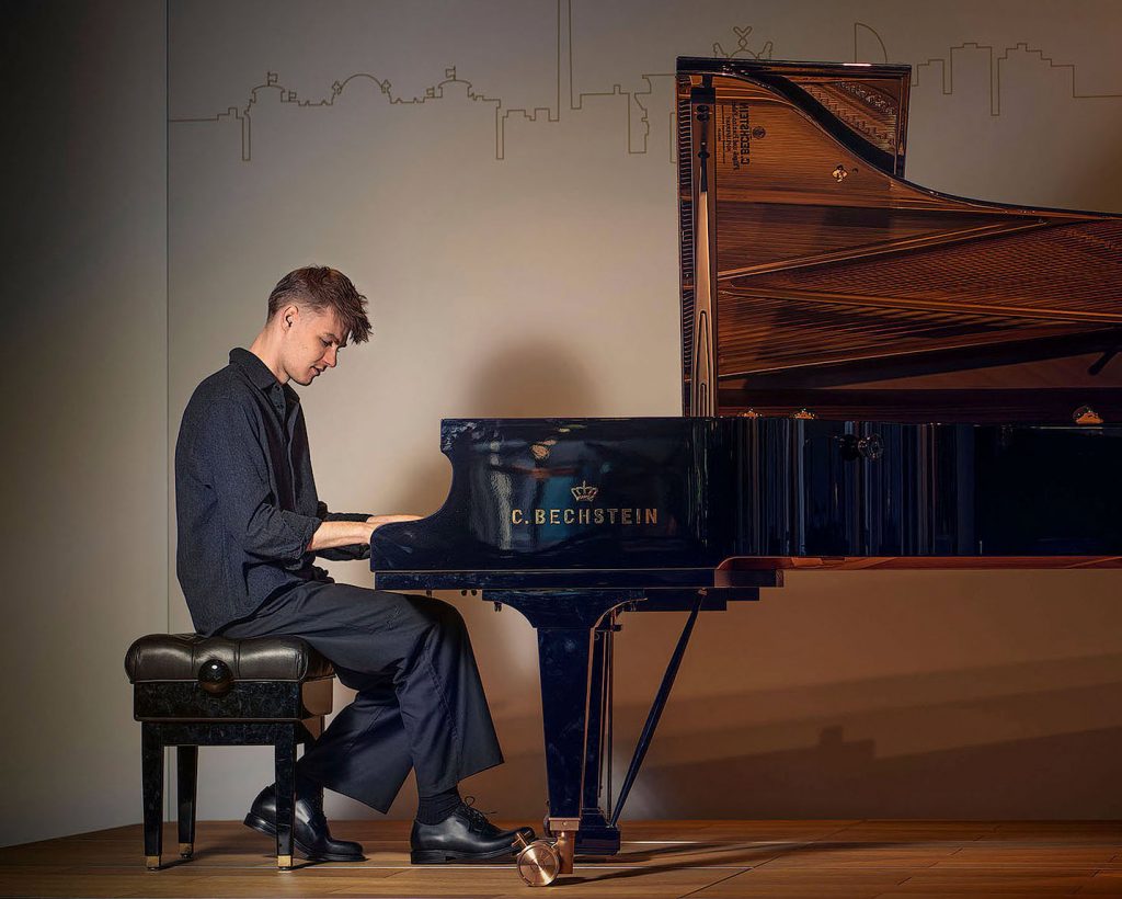 Atmosphärisches Künstlerporträt des Pianisten Janick Čech in einer dunklen Kulisse. Er spielt an einem Flügel, der durch gezielte Beleuchtung hell erstrahlt und im starken Kontrast zur tiefen, schattigen Umgebung steht. Die geheimnisvolle und fokussierte Lichtstimmung betont die Verbindung zwischen Musiker und Instrument.