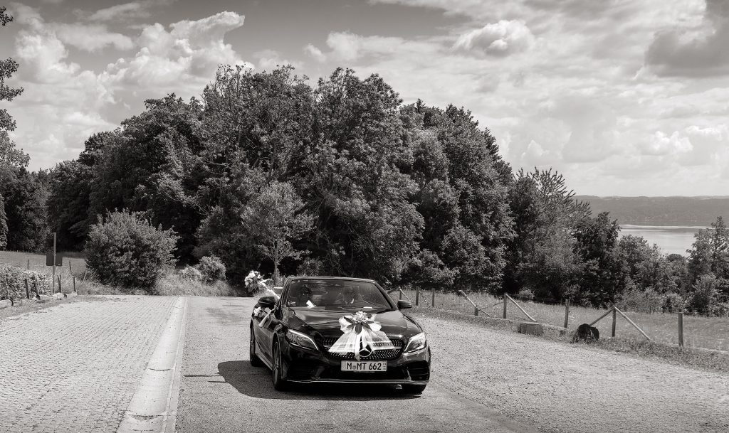 Schwarz-Weiß-Fotografie: Ein offenes Mercedes-Cabriolet als Hochzeitsauto fährt auf einer Landstraße und ist auf der Motorhaube mit weißem Schleifen- und Blumenschmuck dekoriert. Hinter der Frontscheibe ist undeutlich ein Paar zu erkennen, während sie ihre Hand aus dem offenen Verdeck streckt und einen Blumenstrauß hochnimmt. Im Hintergrund sind Büsche, wolkiger Himmel sowie in der Ferne rechts ein See zu sehen.