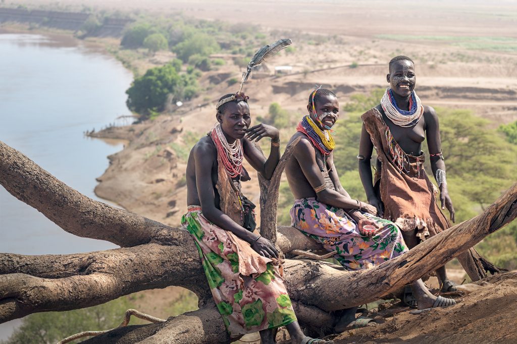 Drei Frauen vom Stamm der Karo mit traditionellem Schmuck und bemaltem Gesicht sitzen auf einem Stamm vor der Kulisse des Omo-Flusses, der sich in der Ferne durch die afrikanische Landschaft schlängelt.