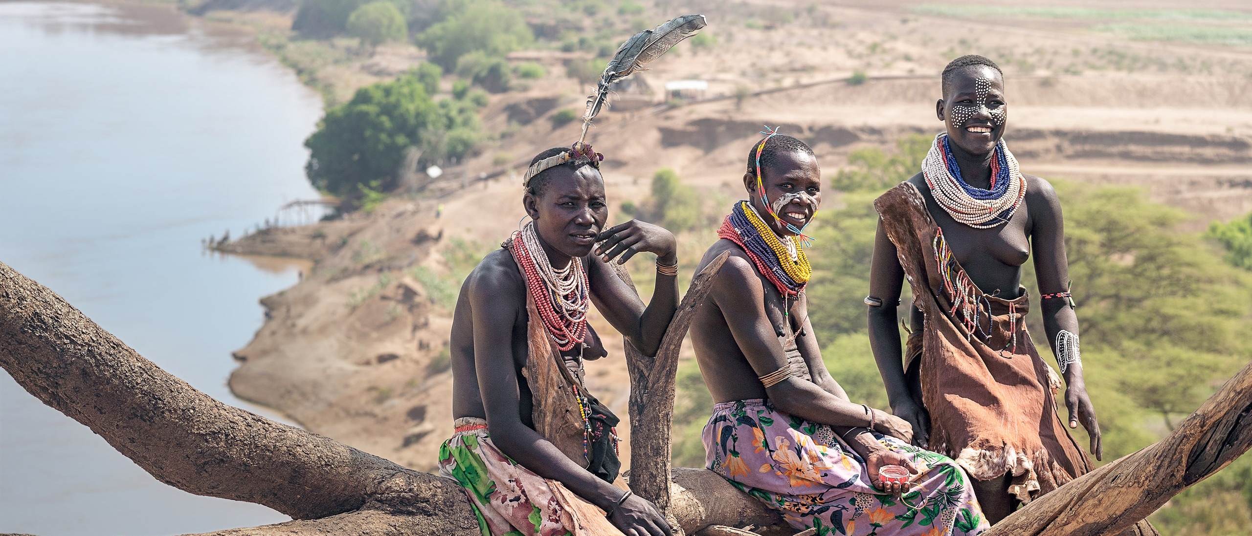 Drei Frauen vom Stamm der Karo mit traditionellem Schmuck und bemaltem Gesicht sitzen auf einem Stamm vor der Kulisse des Omo-Flusses, der sich in der Ferne durch die afrikanische Landschaft schlängelt.