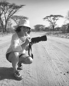Die Fotografin Barbara Westphalen sitzt mit Sonnenhut, Shorts und Turnschuhen auf einer staubigen Sandstraße im äthiopischen Busch und hält eine Kamera mit einer langen Zoomlinse in den Händen.

