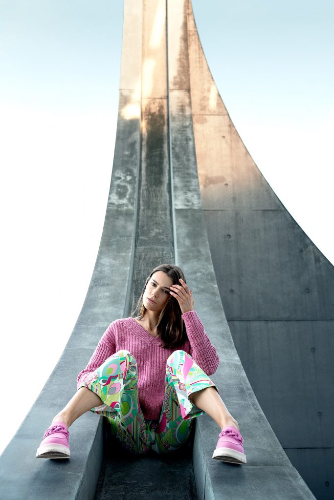 Minimalistisches Fashion-Porträt vor einer Kulisse moderner Architektur mit klaren Linien und Betonstrukturen. Ein Modell sitzt entspannt auf einem architektonischen Element und trägt einen auffälligen, groben pinken Pullover im Kontrast zu einer Hose mit markantem, grafischem Muster.