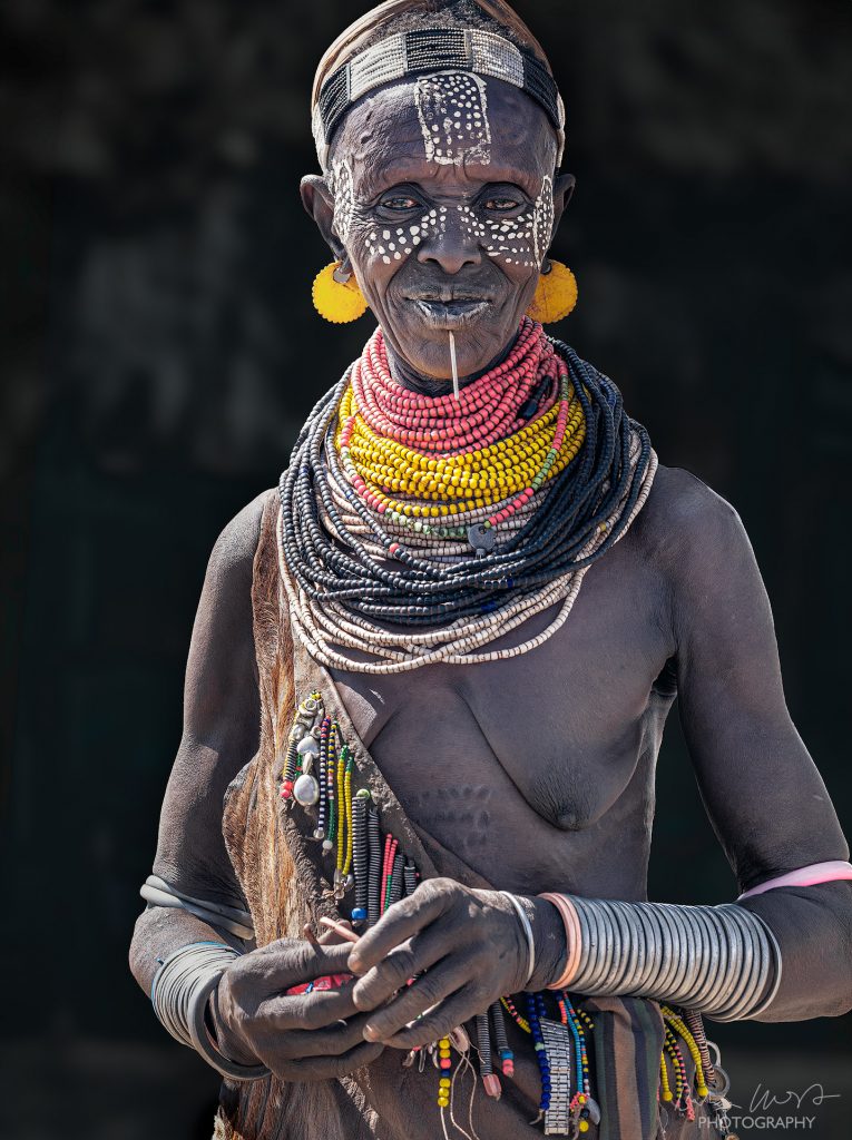 Ein Portrait einer älteren Frau vom Stamm der Karo in Äthiopien vor sehr dunklem Hintergrund. Die Frau hat nicht nur die traditionelle, weiße Bemalung im Gesicht sondern auch einen langen Nagel durchs Kinn gestochen und ein Perlen Stirnband um den Kopf. Ihr Hals wird von unzähligen Lagen Ketten und ihre arme von vielen Lagen Armreifen geschmückt. Um die Schulter trägt sie eine Art Leder Umhang der ebenfalls mit Perlen dekoriert ist. Am Bauch sieht man Schmucknarben.