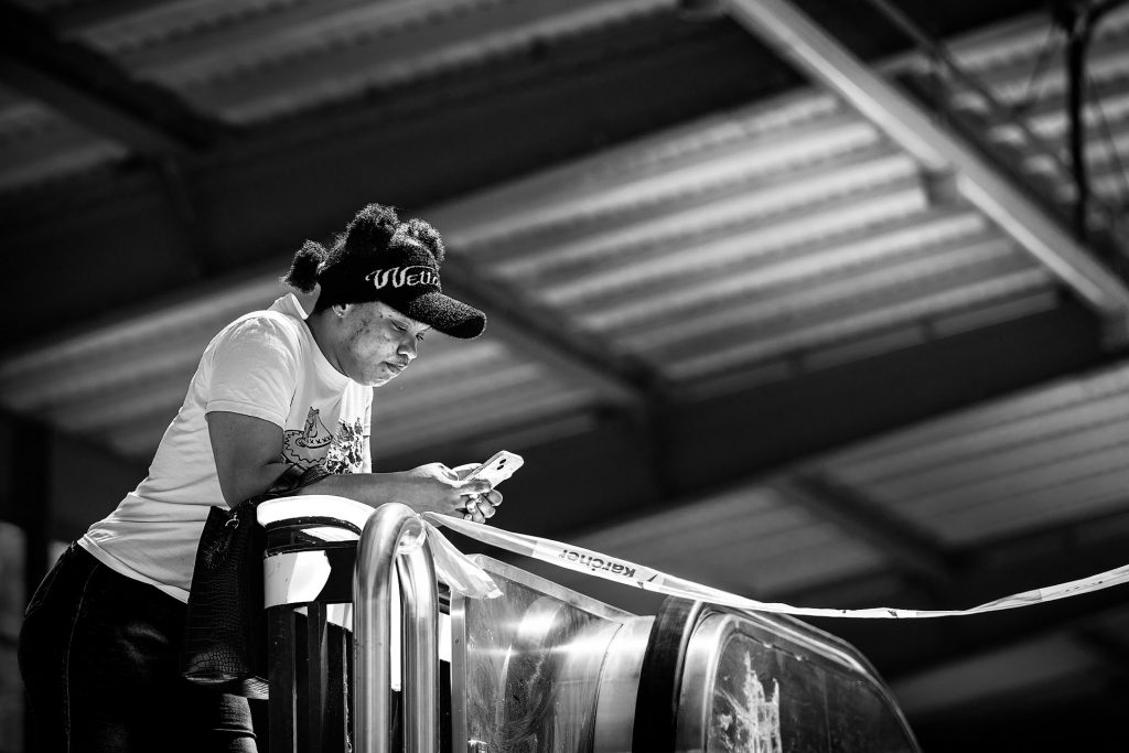 Schwarz-Weiß Fine-Art Streetfotografie einer seitlich aufgenommenen Frau mit dunkler Hautfarbe, bekleidet mit Jeans, weißem T-Shirt und einer Wollmütze. Sie stützt sich mit den Ellenbogen auf das Geländer einer Rolltreppe und bedient ihr Mobiltelefon. Quer vor der Rolltreppe verläuft ein rot-weißes Absperrband.