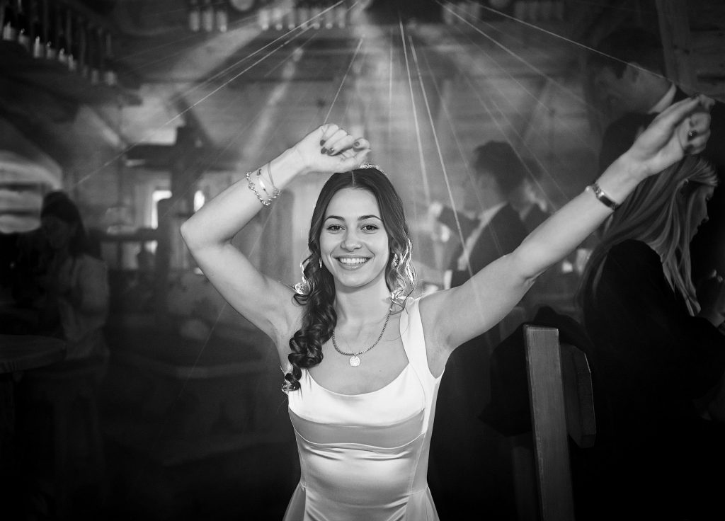 Schwarz-Weiß-Fotografie: Foto einer jungen, dunkelhaarigen Frau mit langen Locken. Sie lacht in die Kamera, hat die Arme weit nach oben ausgestreckt und trägt ein weißes, abendliches Kleid. Im Hintergrund eine Partyszene.