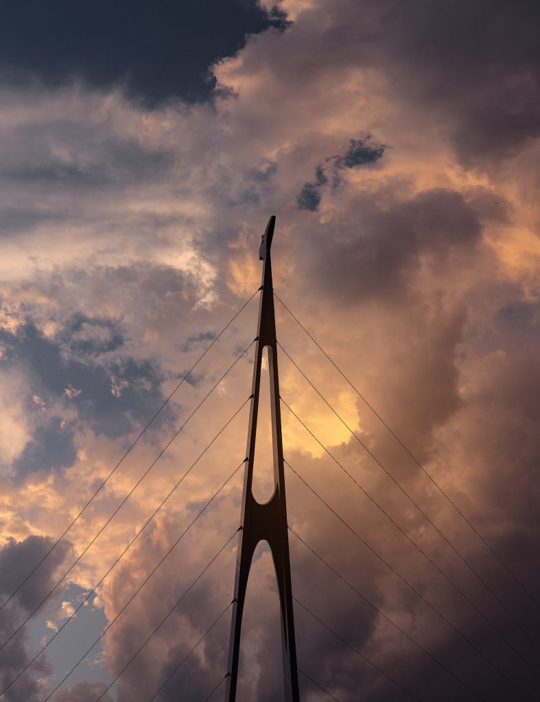 Der obere Teil eines Brückenpfeilers der Fußgängerbrücke in Kuching ist zu sehen. Schwarze Seilaufhängungen verlaufen diagonal zu den unteren Bildschirmrändern. Im Hintergrund erstreckt sich ein dramatisch rot gefärbter Abendhimmel mit malerischen, kräftigen Wolken.