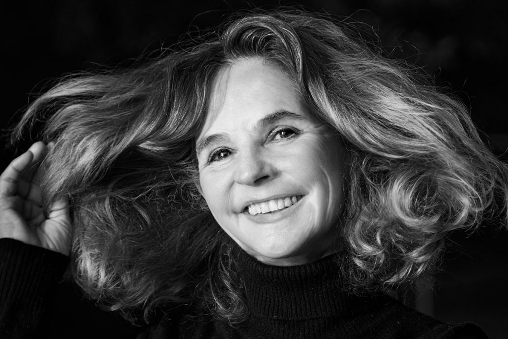 Schwarz-Weiß-Porträt der Fotografin Barbara Westphalen. Auf dem Bild ist nur ihr Kopf zu sehen. Sie wirft ihre üppigen dunklen, lockigen Haare zur Seite und lächelt vor einem dunklen Hintergrund direkt in die Kamera.